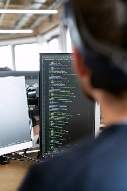 The image depicts a close-up view of a person working in an indoor office environment, focusing on a computer monitor displaying lines of code in various colors. The individual is seated at a desk wit