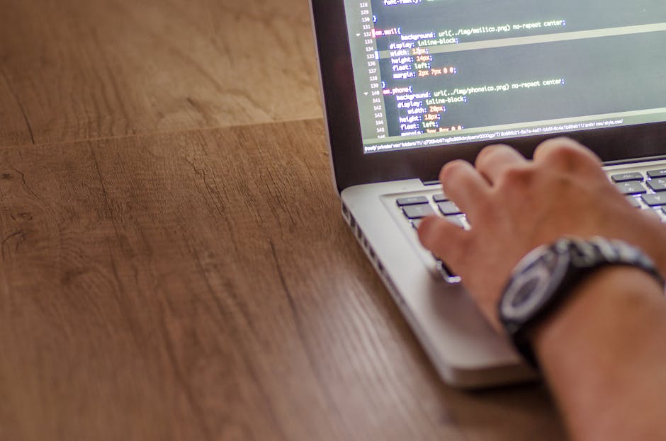 The image depicts a close-up view of a person working in an indoor office environment, focusing on a computer monitor displaying lines of code in various colors. The individual is seated at a desk wit